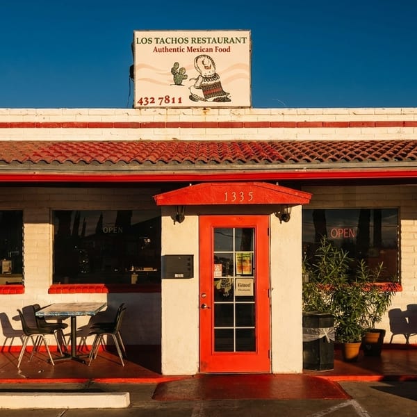 Exterior of Los Tachos Restaurant with a red door, outdoor seating, and a sign against a blue sky.