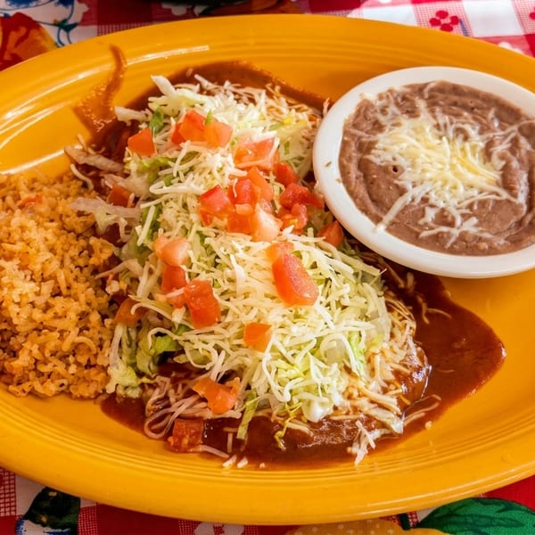 A vibrant yellow plate holds enchiladas topped with lettuce, tomatoes, and cheese, alongside rice and refried beans.