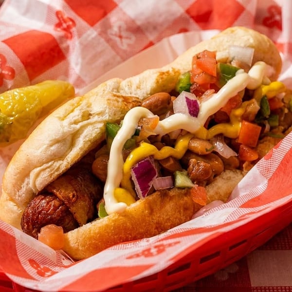 Bacon-wrapped hot dog with beans, diced vegetables, mustard, and mayonnaise in a red basket.