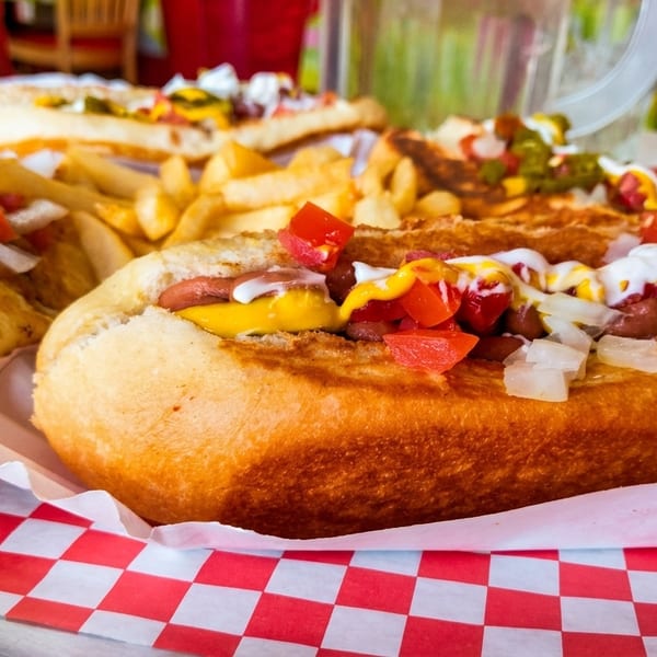 Three loaded hot dogs with fries, mustard, ketchup, onions, and tomatoes on a red checkered tray.