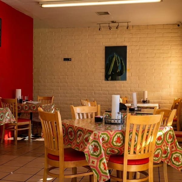A brightly lit restaurant interior with red and brick walls, wooden chairs, and colorful patterned tablecloths.
